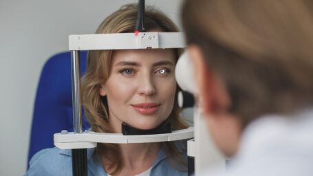 Portrait of smiling female client checking eyesight with equipment opposite doctor. Ophthalmology concept